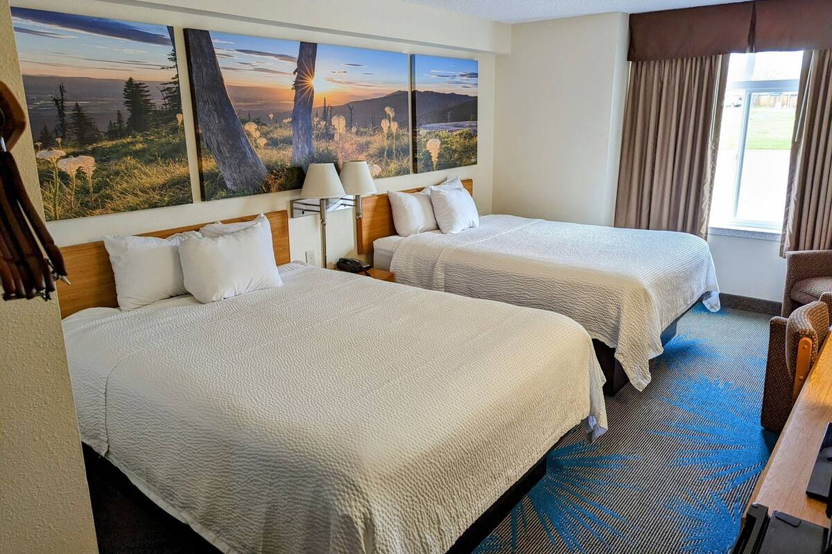 A hotel room with two beds, a large window with curtains, a painting of a landscape, and a desk with a chair.