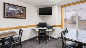 A hotel room with a desk, computer, and two chairs. There is a television mounted on the wall and a window with blinds.