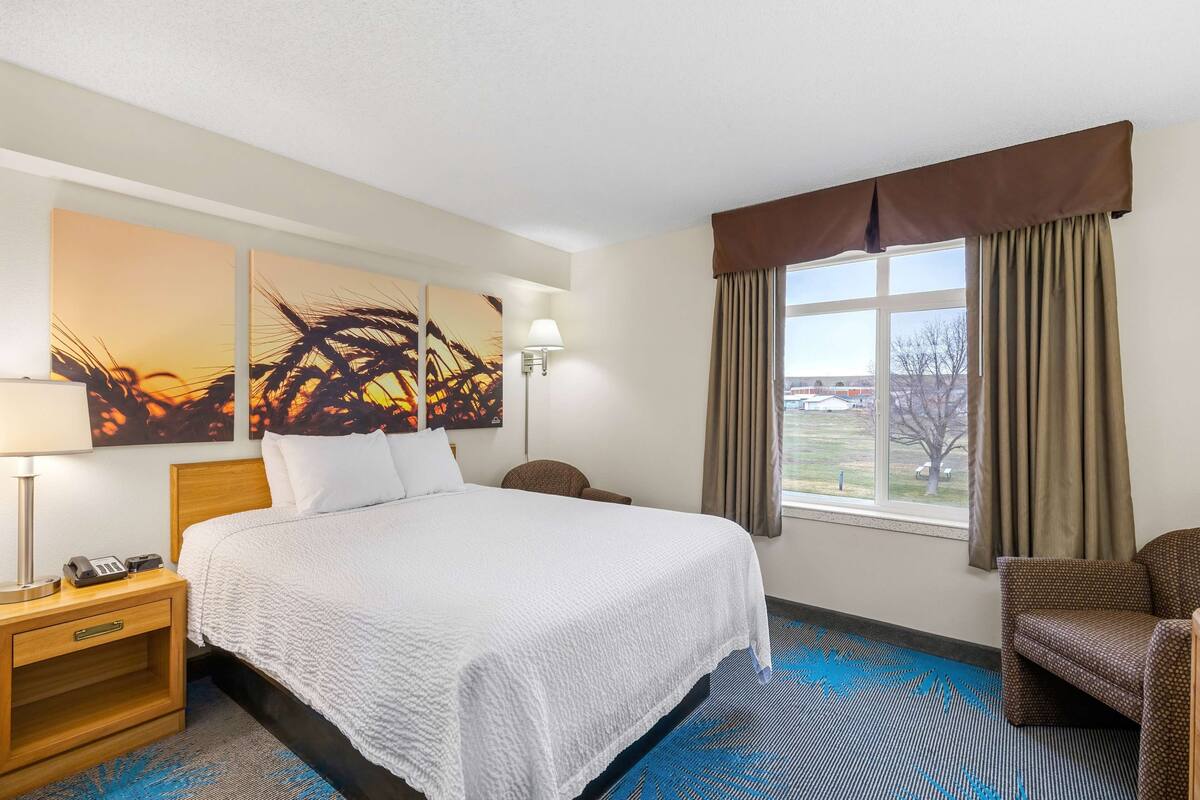 A hotel room with a large bed, a nightstand with a lamp, a chair, and a window with curtains.
