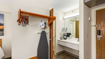 A hotel room with a wooden ironing board, a mirror, a sink with toiletries, and a door with a lock.