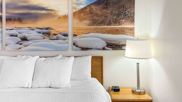 A hotel room with a bed, white pillows, a nightstand with a lamp, and a painting of a snowy landscape.