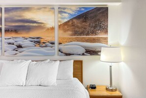 Une chambre d’hôtel avec un lit, des oreillers blancs, une table de chevet avec une lampe et un tableau représentant un paysage enneigé.
