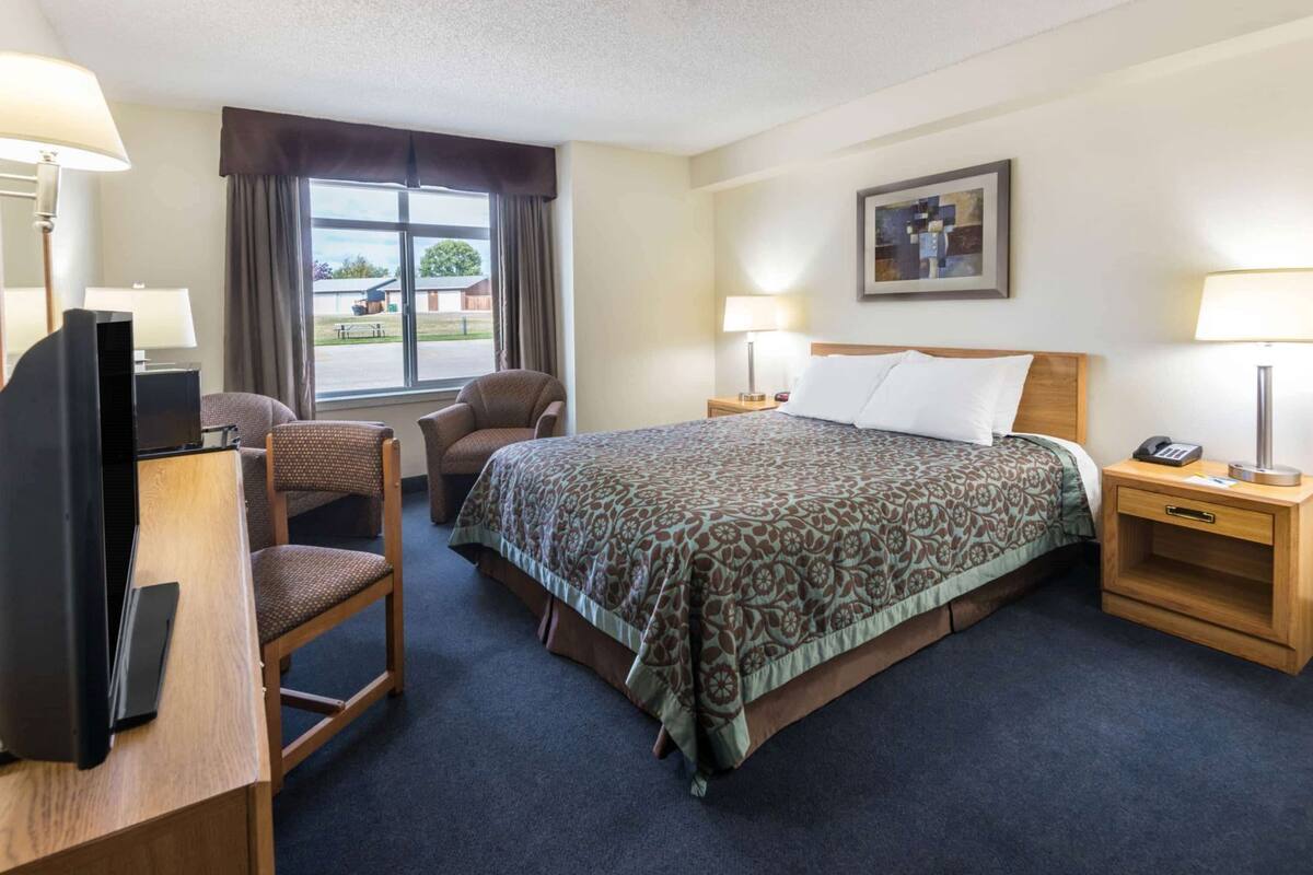 A hotel room with a bed, two chairs, a desk with a TV, bedside tables with lamps, and a window with a view of the outside.