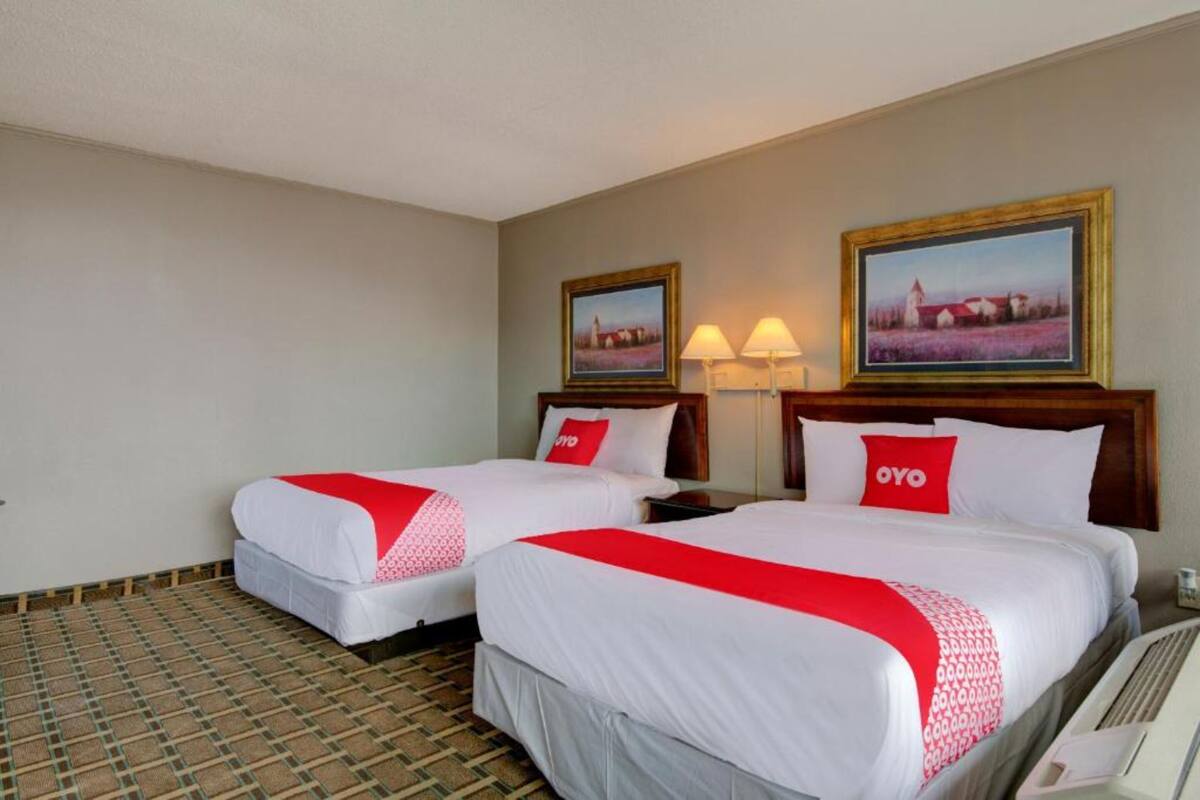 A hotel room with two beds, each with a red and white bedspread, a wooden headboard, and a nightstand with a lamp. There are two framed pictures on the wall, one depicting a rural scene and the other a field of flowers.