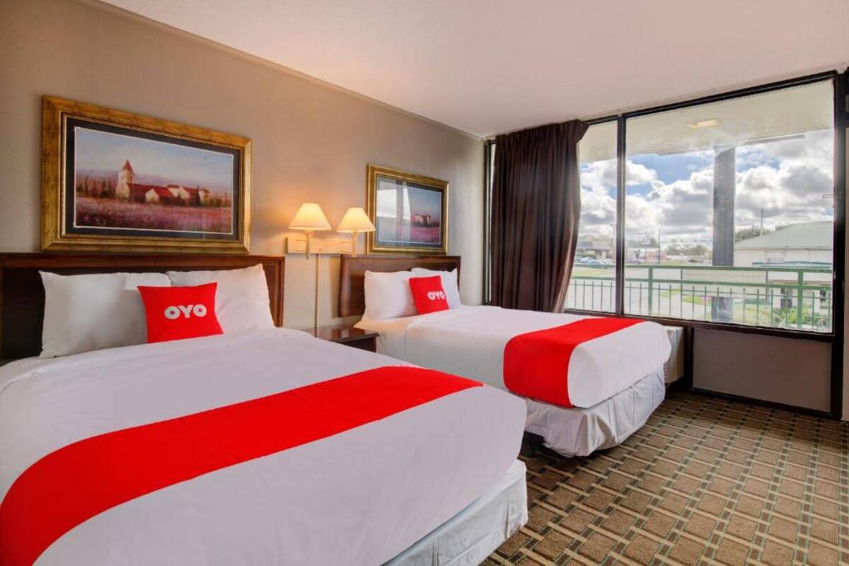 A hotel room with two beds, each with a red and white bedspread, a large window with a view of a cityscape, and two wall-mounted lamps.