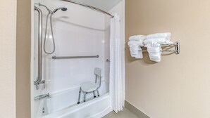 A bathroom with a walk-in shower, grab bars, a small chair, and a towel rack. - Quality Inn West Plano - Dallas (Plano)