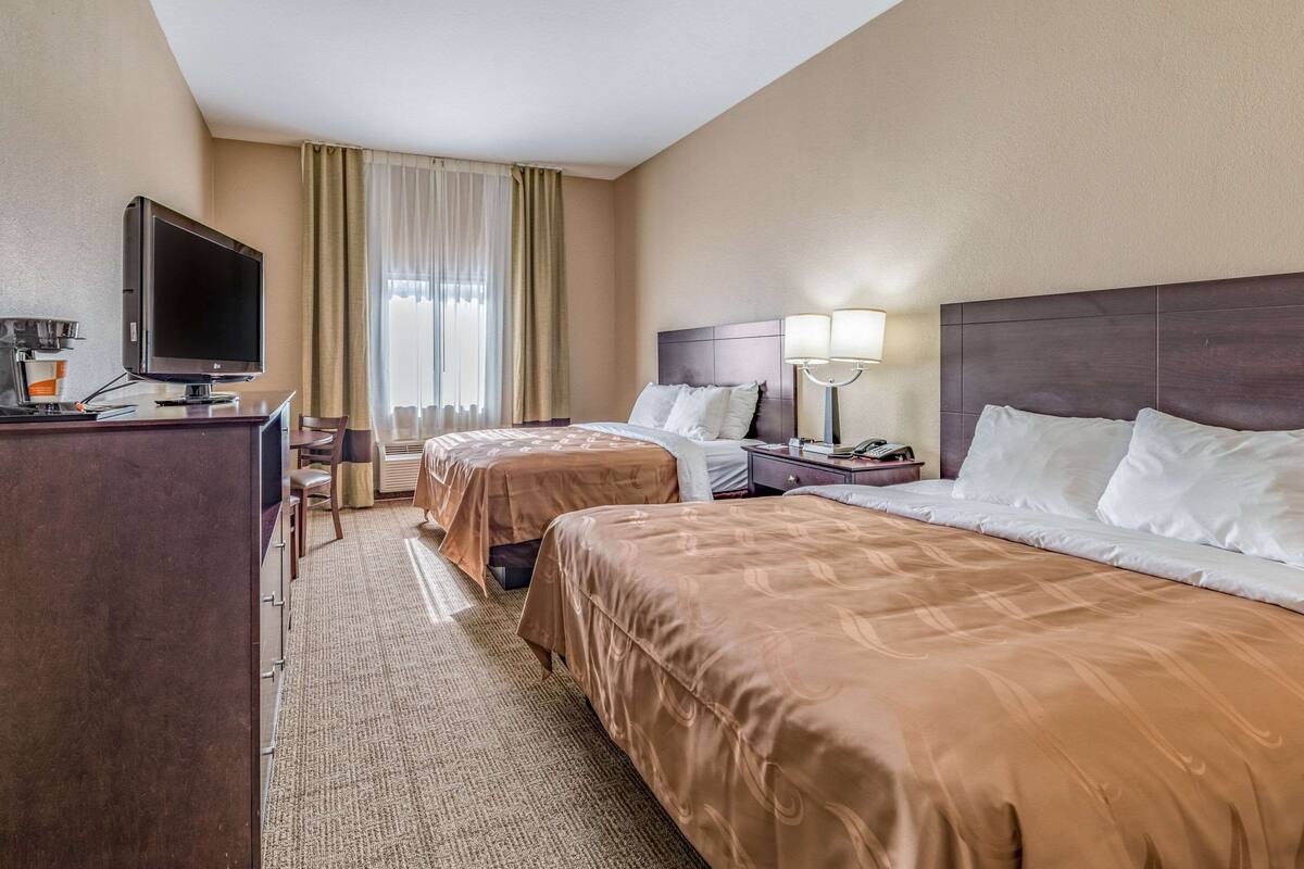 A hotel room with two beds, a desk with a TV, a lamp, and a window with curtains.
