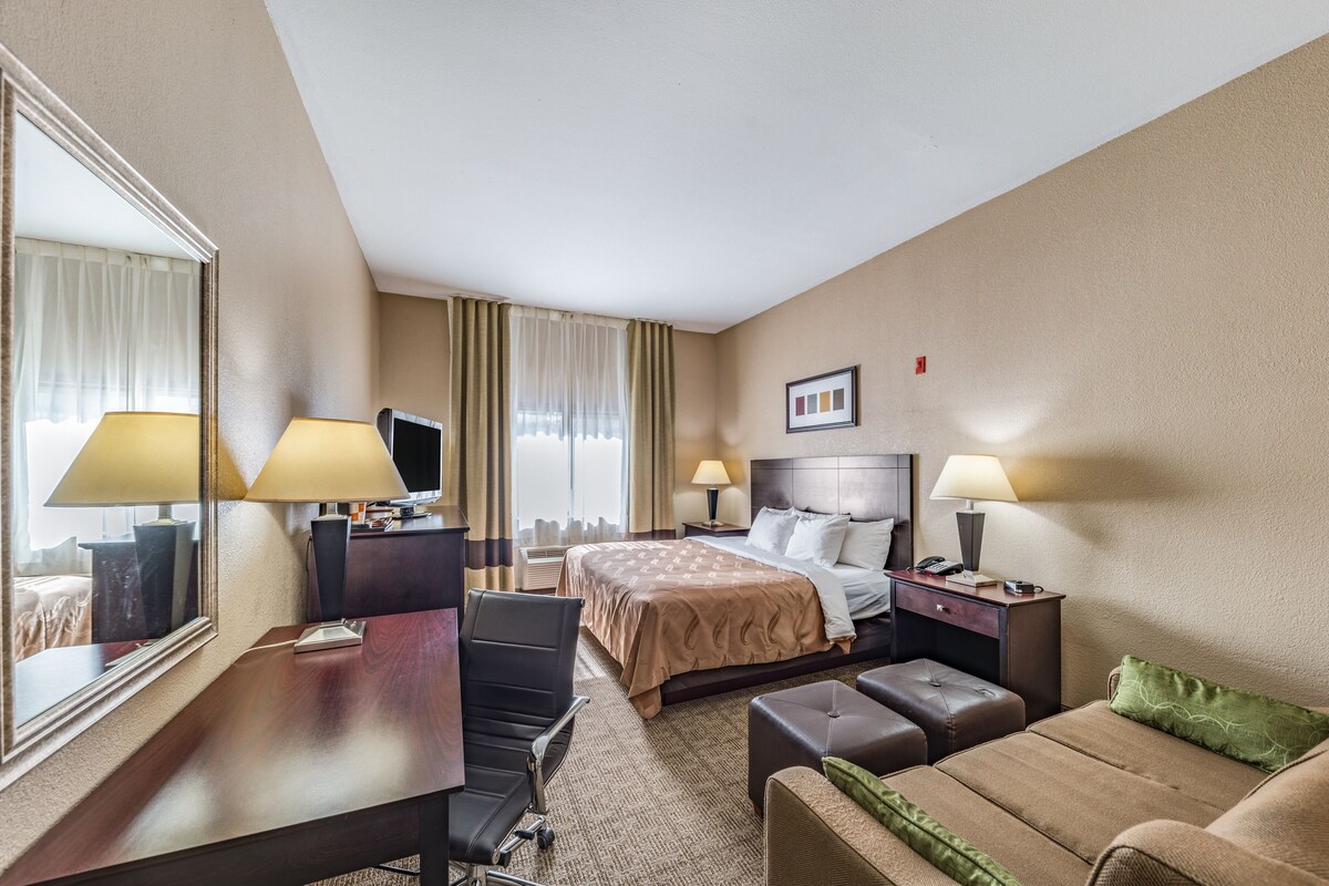 A hotel room with a bed, two bedside lamps, a desk with a chair, a television, and a large mirror.