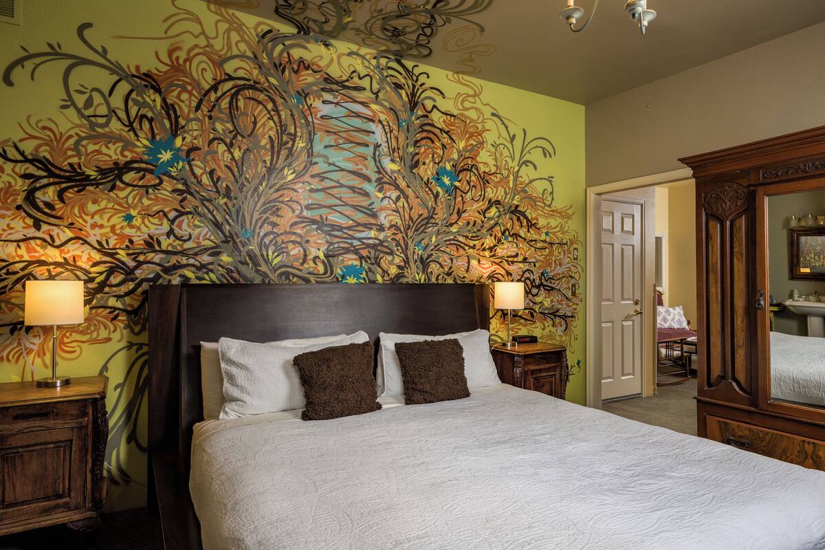Une chambre d’hôtel avec un grand lit, deux tables de chevet, une tête de lit en bois et une fresque florale sur le mur.