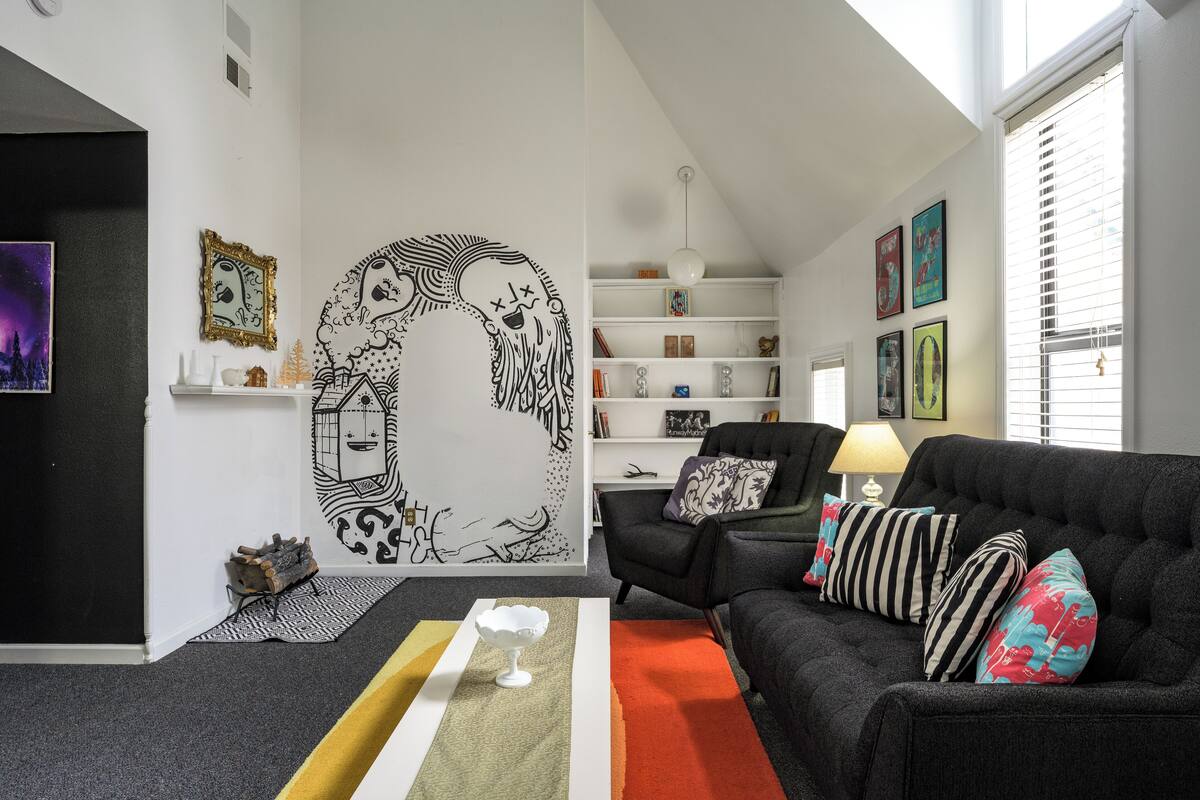 Un salon moderne avec un canapé noir, des coussins colorés, une table basse blanche et une grande fresque murale.