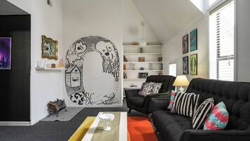 A modern living room with a black sofa, colorful pillows, a white coffee table, and a large wall mural.