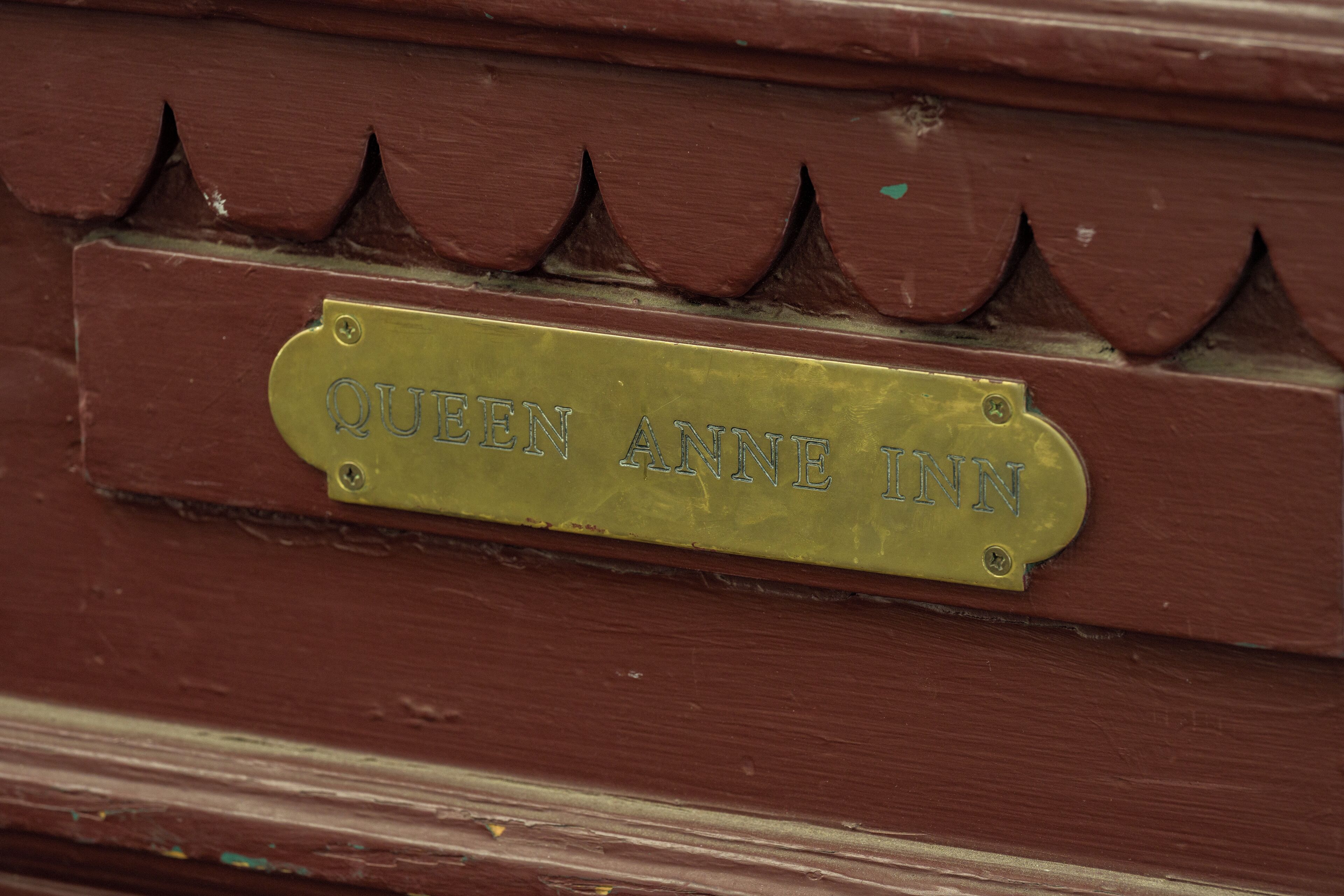 A brass plaque with the inscription "QUEEN ANNE INN" on a wooden surface with a decorative pattern.