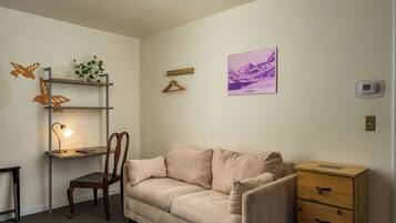A room with a beige sofa, a wooden desk with a lamp, a chair, a small wooden cabinet, a wall-mounted shelf with a plant, and a framed picture.