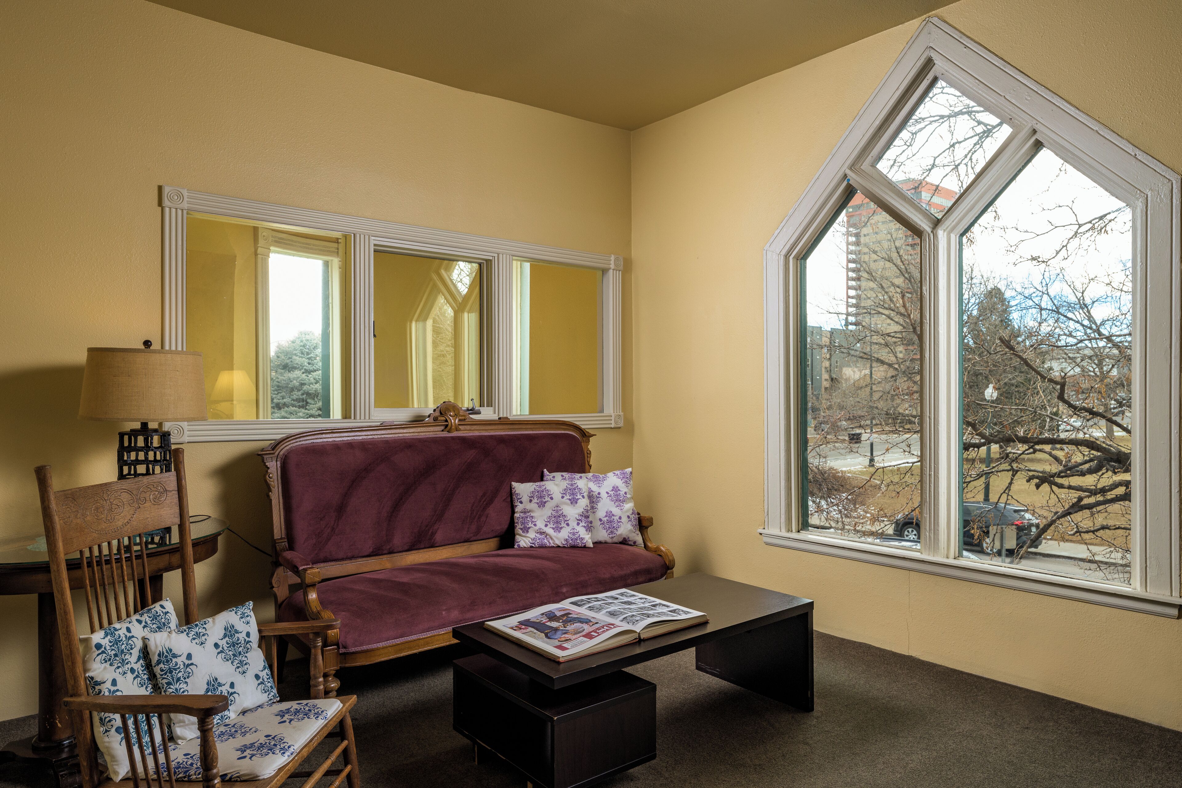 A room with a wooden chair, a sofa, a table with a magazine, and a large window with a view of trees and buildings.