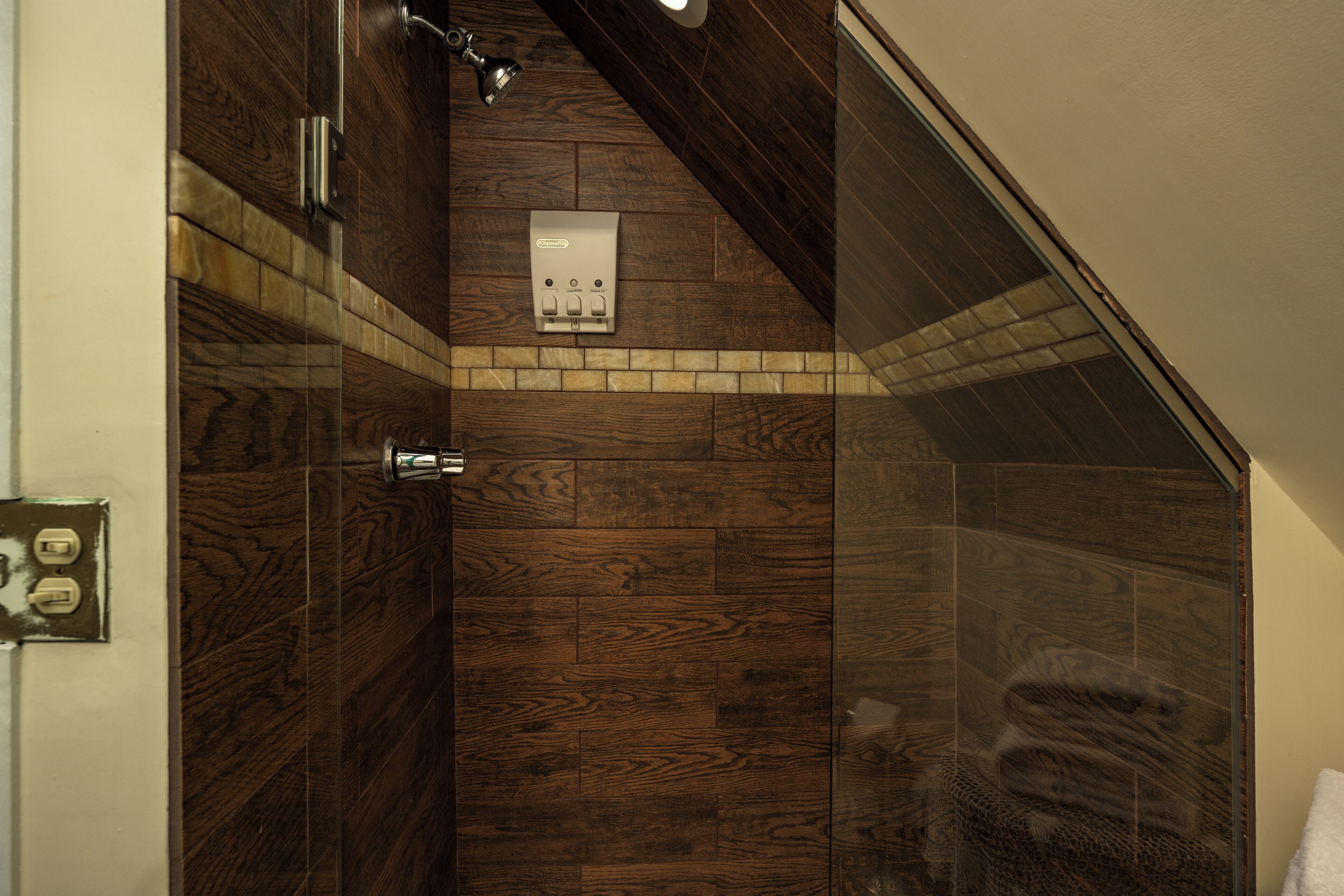 Ducha con suelo de madera y puerta de cristal, una lámpara sobre ella y un panel de control en la pared.