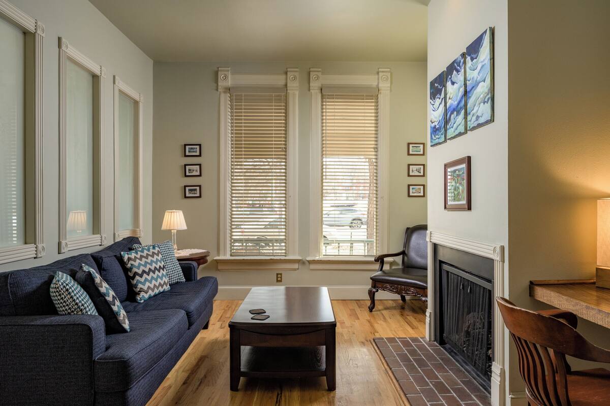 Un salon avec un canapé bleu foncé, une table basse en bois et une cheminée. Il y a trois fenêtres équipées de stores, et une chaise est placée près de la cheminée.
