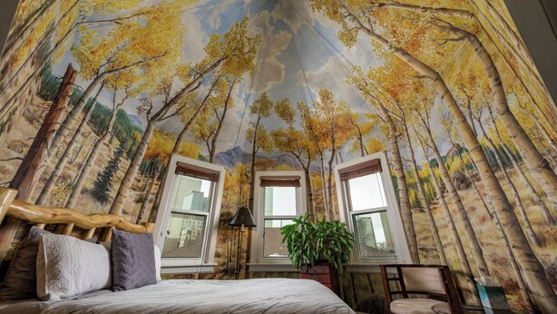 A bedroom with a mural of a forest canopy, a bed with pillows, a nightstand, a lamp, and a window with a view of buildings. - Queen Anne Bed And Breakfast (Denver)