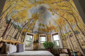 Une chambre avec une fresque représentant la canopée d’une forêt, un lit avec des oreillers, une table de chevet, une lampe et une fenêtre donnant sur des immeubles.