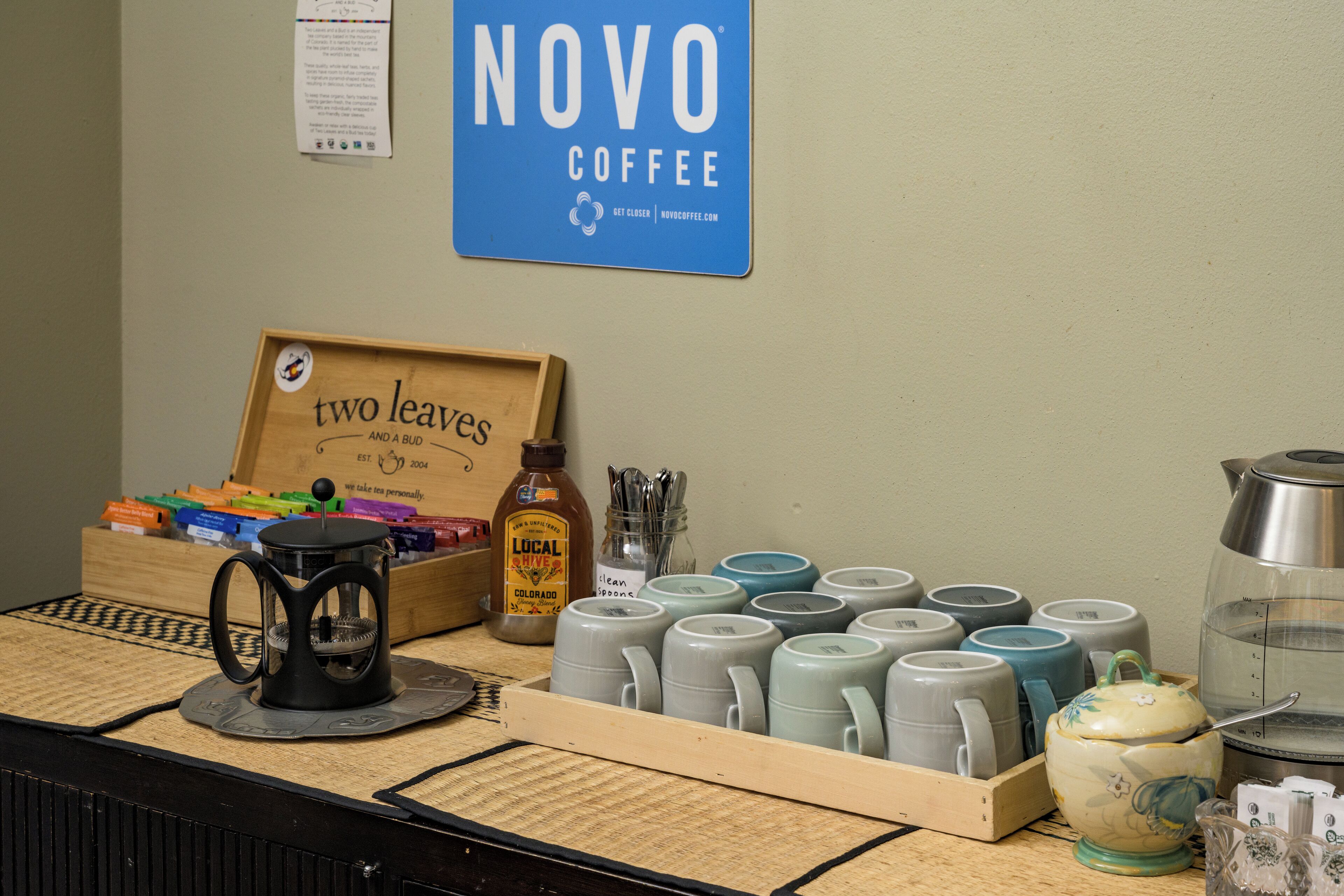Een toonbank van een koffiebar met een bord waarop 'NOVO COFFEE' staat, een koffiezetapparaat, mokken en een houten bordje met de tekst 'two leaves and a bud'.