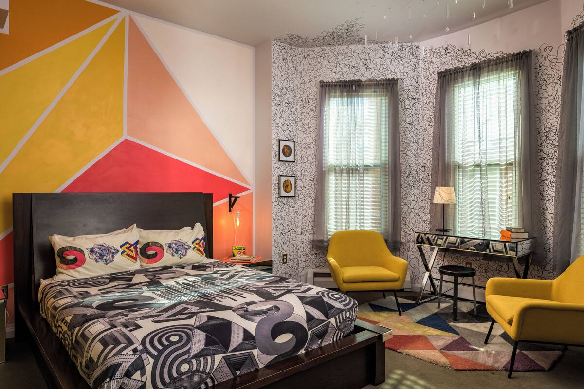 Une chambre à coucher moderne avec une couvre-lit à motifs géométriques, un bureau avec une lampe et deux chaises jaunes.