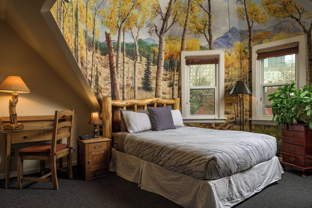Une chambre à coucher comprenant un lit, un bureau, une chaise, une commode et une fenêtre donnant sur des immeubles.
