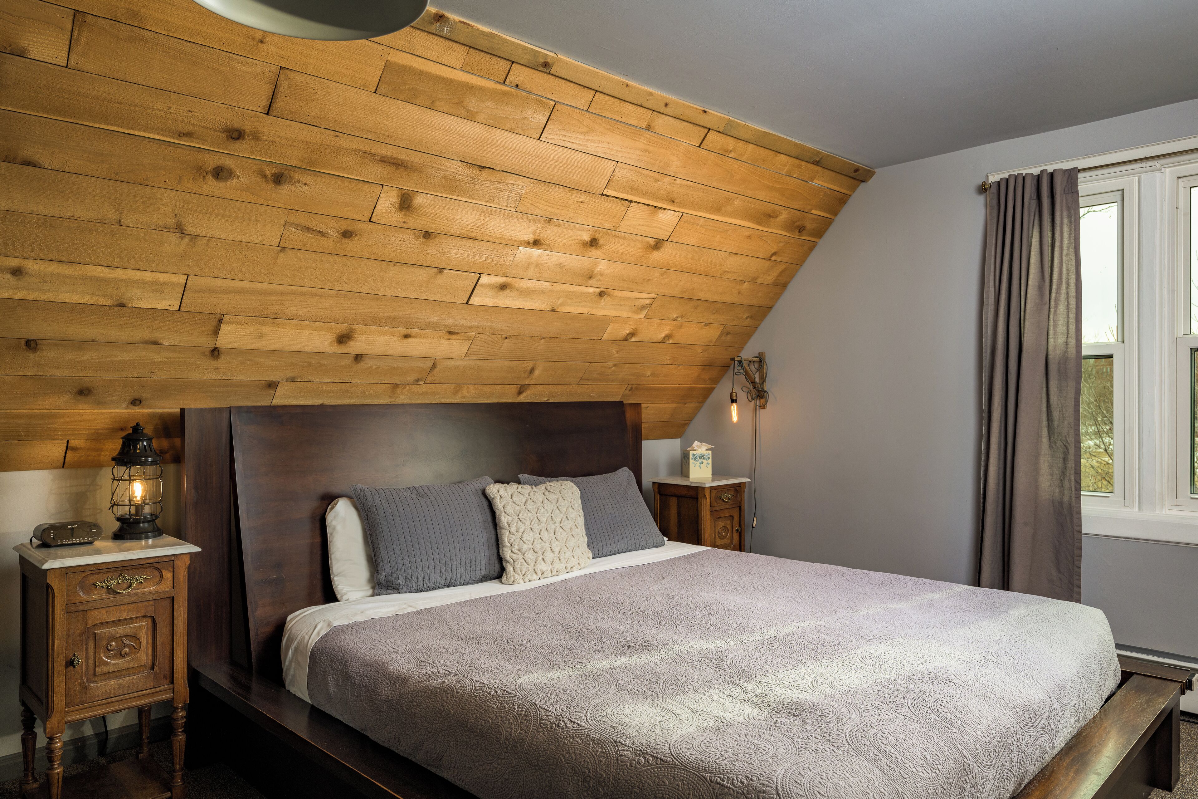 Ein Schlafzimmer mit holzverkleideter Schräge, einem Bett mit gemustertem Überwurf, einem Nachttisch mit Laterne und einem Fenster mit Vorhängen.