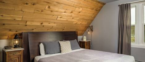 Ein Schlafzimmer mit holzverkleideter Schräge, einem Bett mit gemustertem Überwurf, einem Nachttisch mit Laterne und einem Fenster mit Vorhängen.