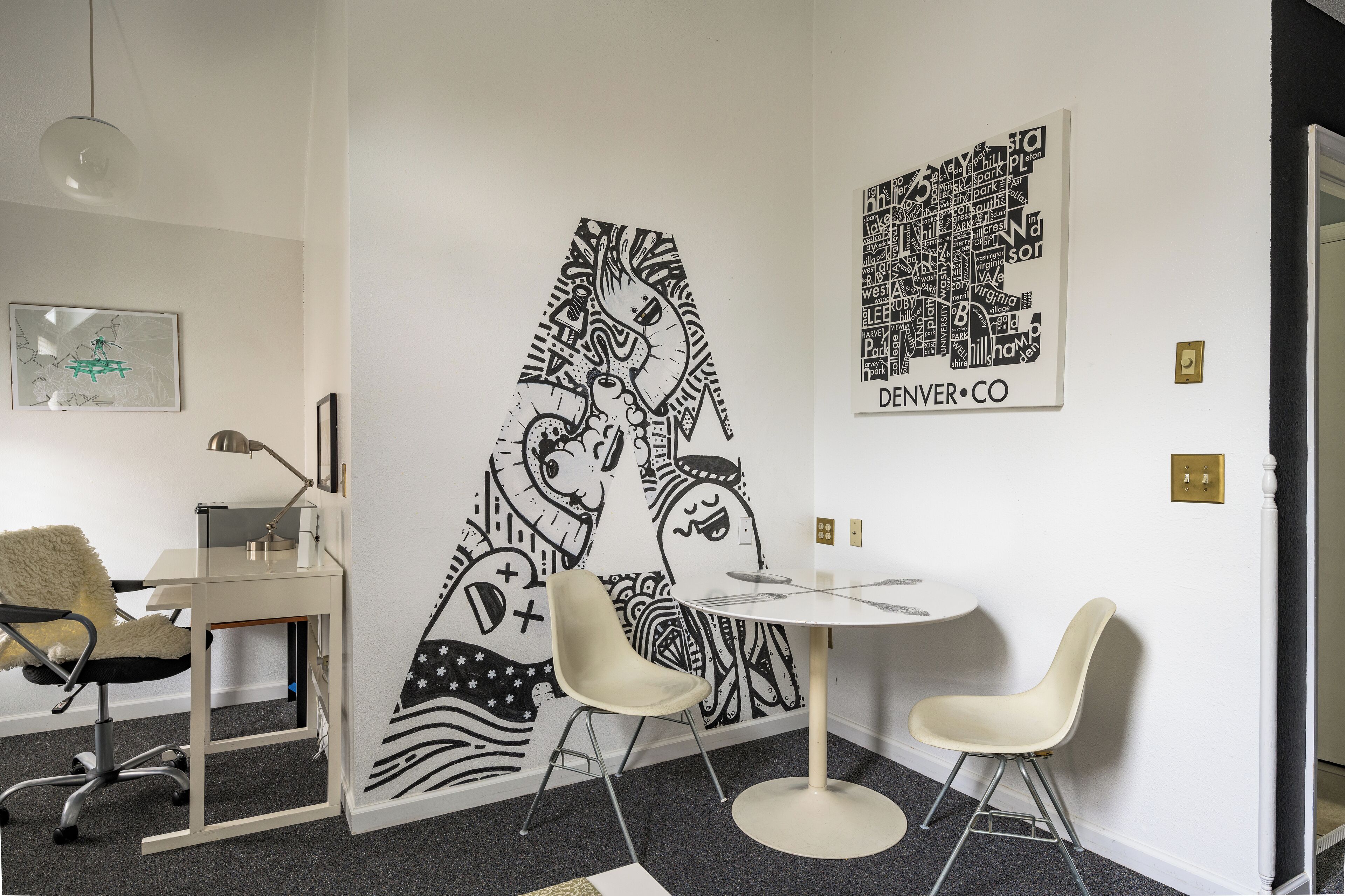 A modern room with a white wall, a large black and white mural, a round table, two white chairs, and a desk with a chair.
