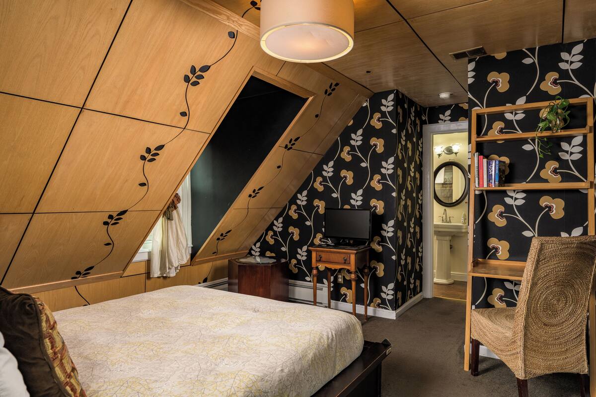 Une chambre avec un plafond en pente, un lit, une chaise en bois, une bibliothèque et un téléviseur.