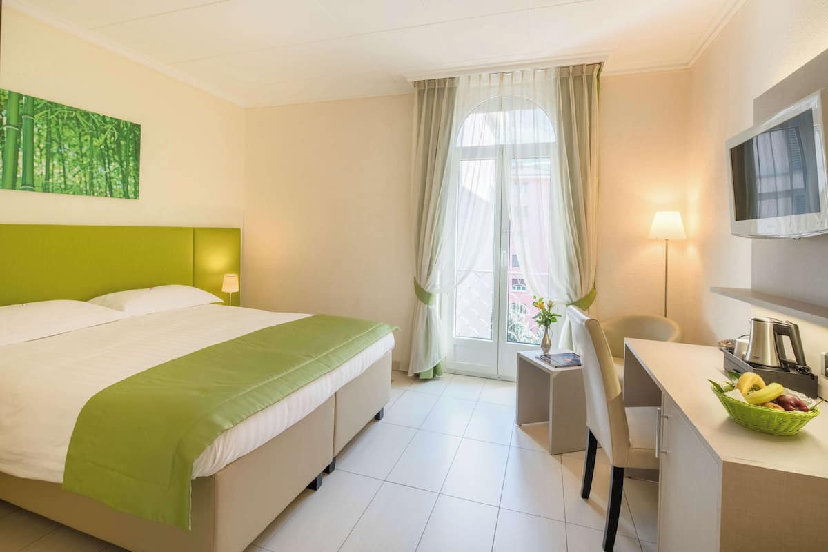 Una habitación de hotel con cama, escritorio con silla, televisor y ventana con cortinas.