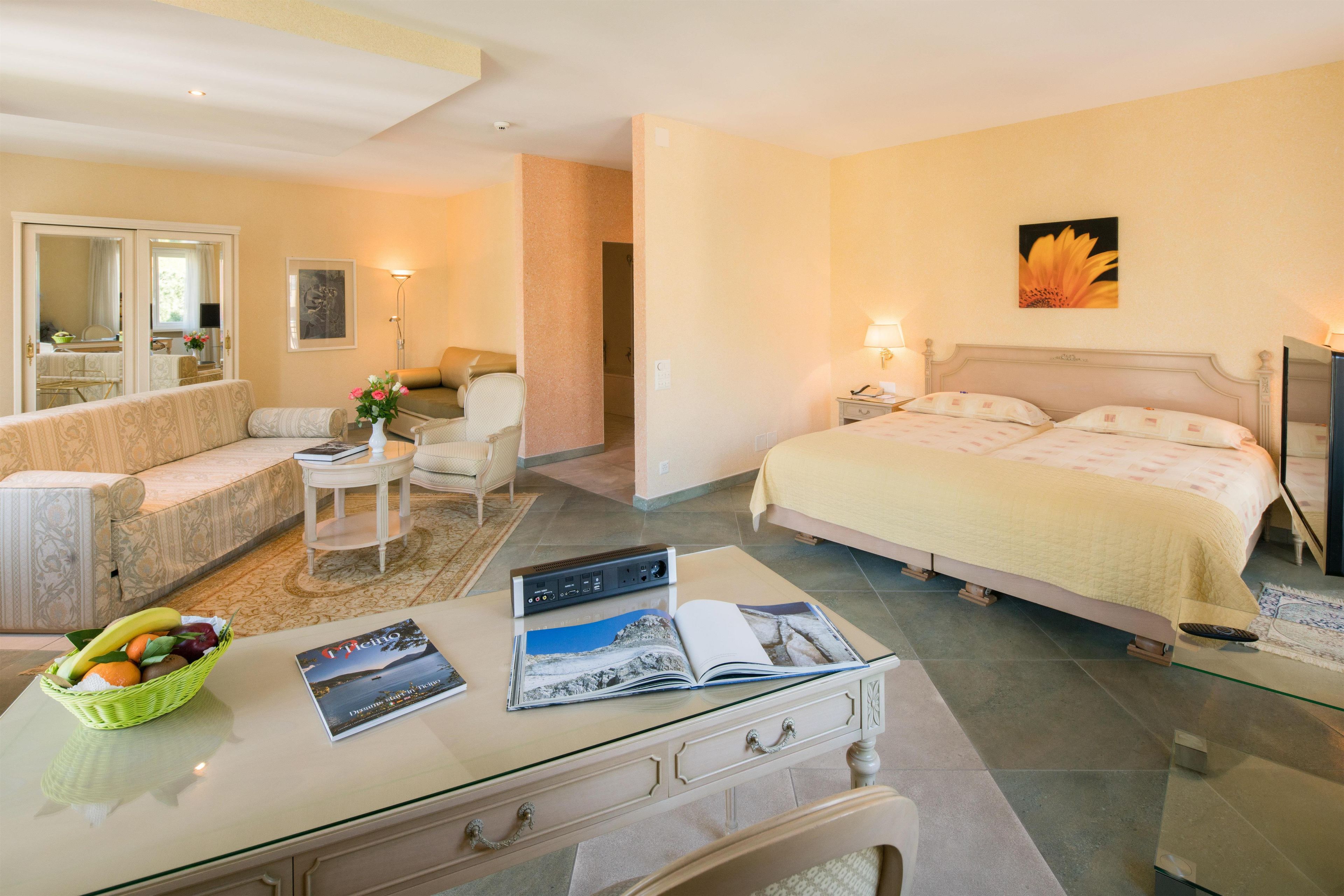 Una habitación de hotel con una cama, un sofá, una mesita con una cesta de frutas y un escritorio con una computadora y revistas.