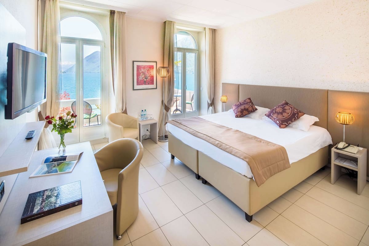 Habitación de hotel con cama, escritorio con silla, televisor, ventana con vistas y mesita de noche con lámpara.