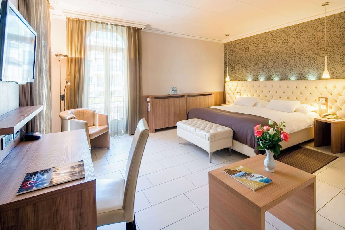 Habitación de hotel con cama, escritorio, silla, mesa pequeña, televisor y ventana con cortinas.