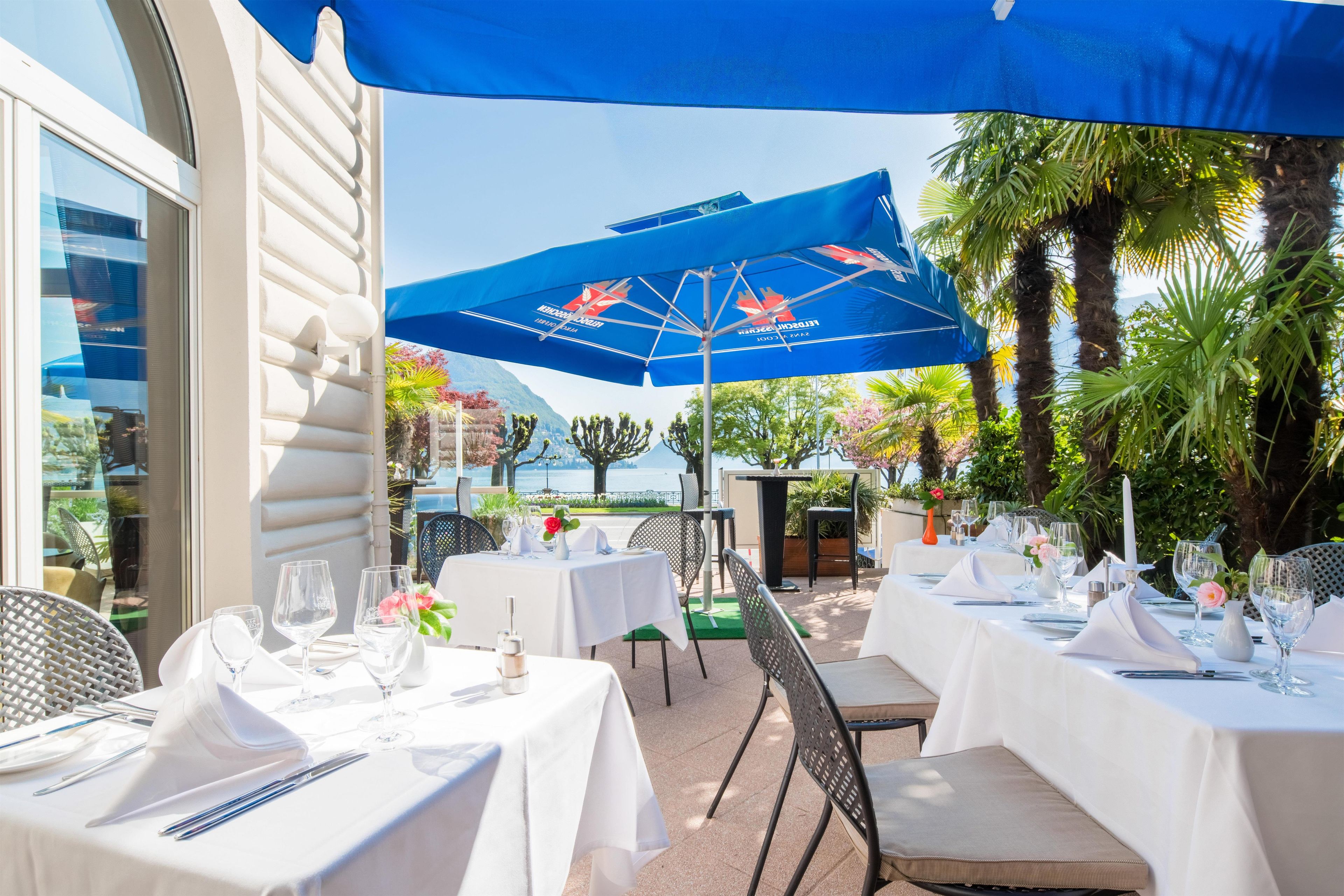 Un área de comedor al aire libre con mesas para dos, cubierta por sombrillas azules, rodeada de follaje y vistas al mar.