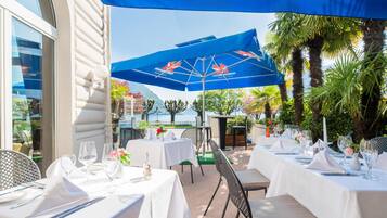 Un área de comedor al aire libre con mesas para dos, cubierta por sombrillas azules, rodeada de follaje y vistas al mar.