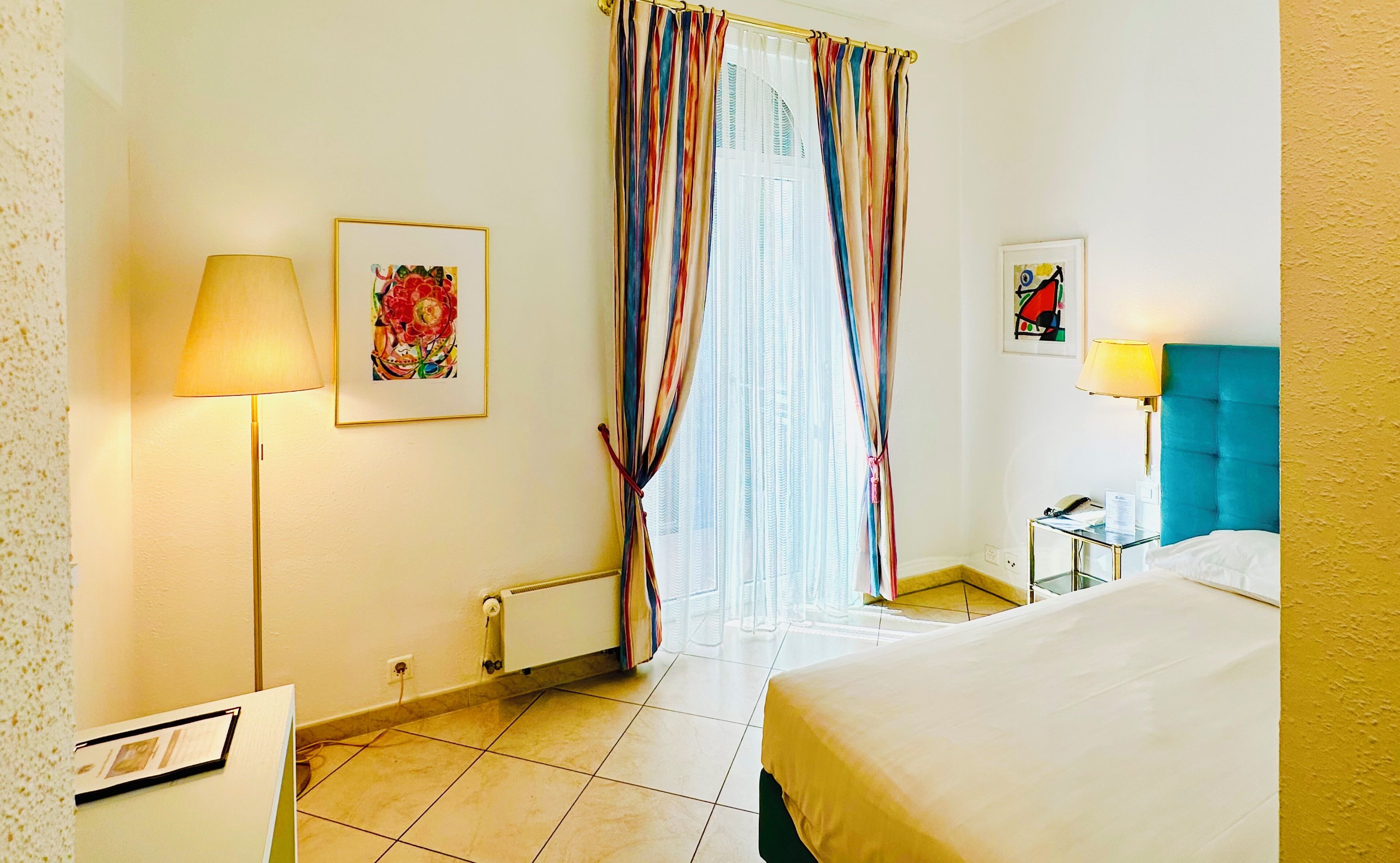 Una habitación de hotel con una cama, un escritorio con lámpara, un radiador y dos cuadros en la pared.