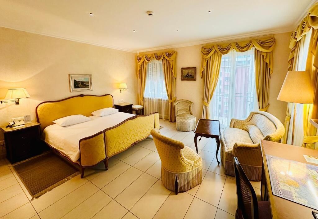 A hotel room with a large bed, a sitting area with a sofa and armchairs, a small table, and two windows with curtains.