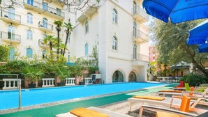 Un área de piscina con sillas de descanso y sombrillas, rodeada por un edificio con varios balcones y vegetación.