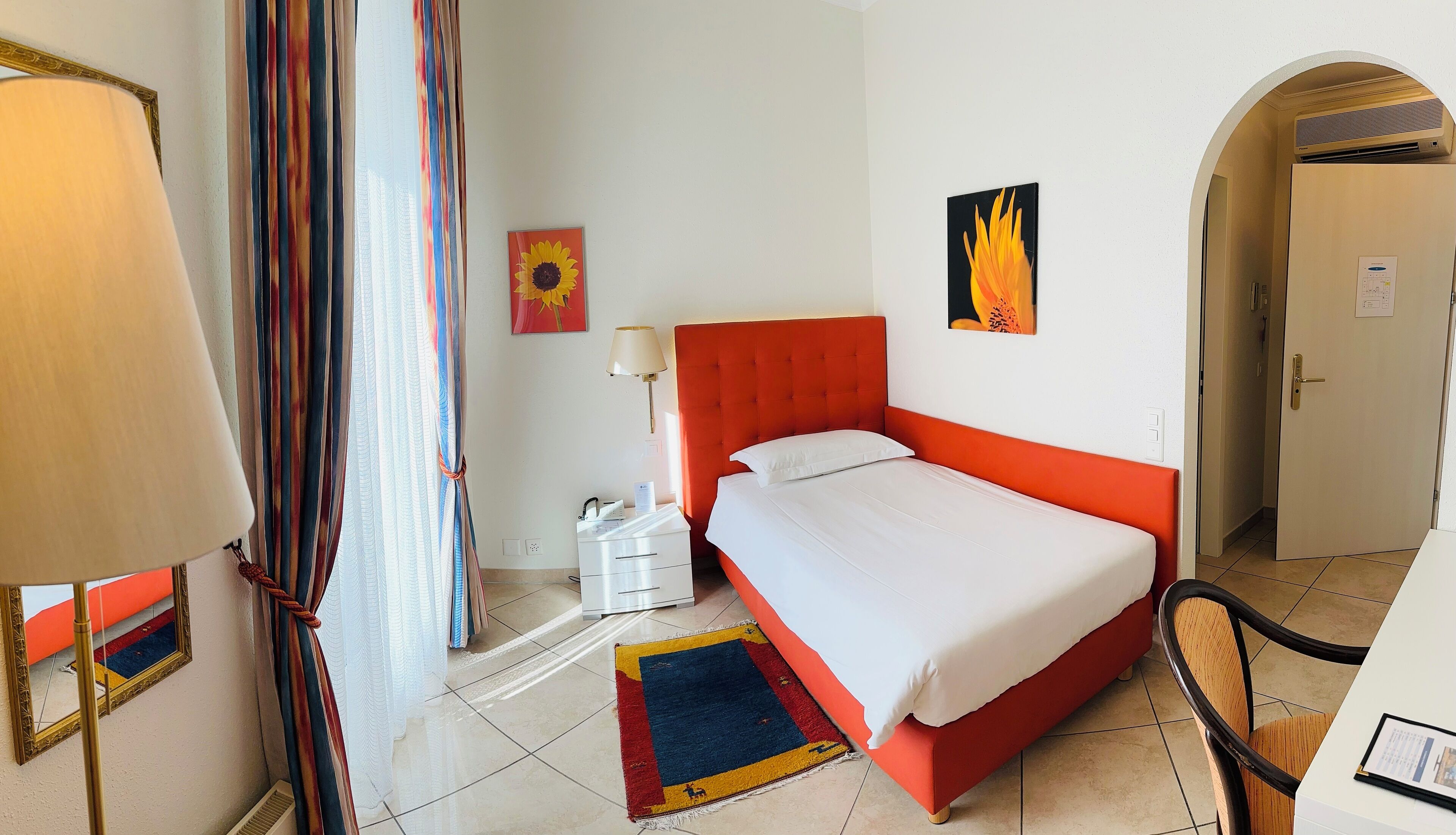 A hotel room with a red upholstered bed, white linens, a colorful rug, a bedside table with a lamp, and two wall-mounted paintings.