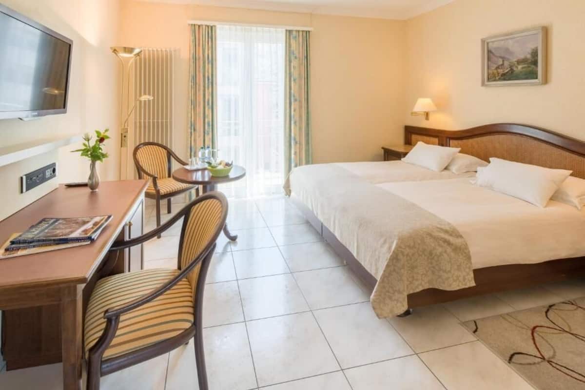 Habitación de hotel con cama, escritorio, sillas, televisor y ventana con cortinas.
