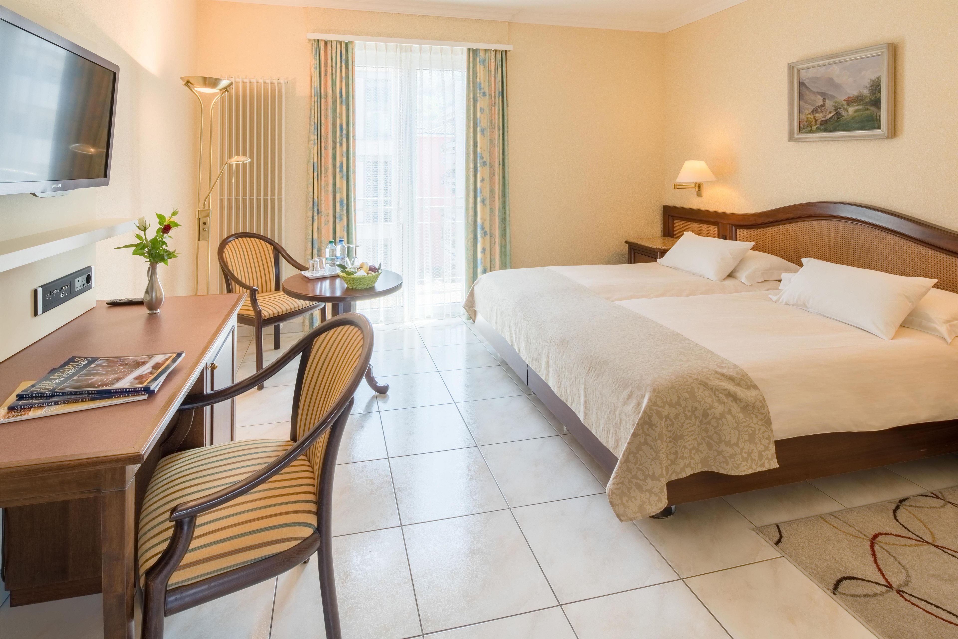 A hotel room with a bed, a desk with chairs, a television, and a painting on the wall.