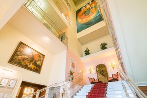 Una majestuosa escalera con alfombra roja que conduce a un rellano con un cuadro grande y un candelabro.