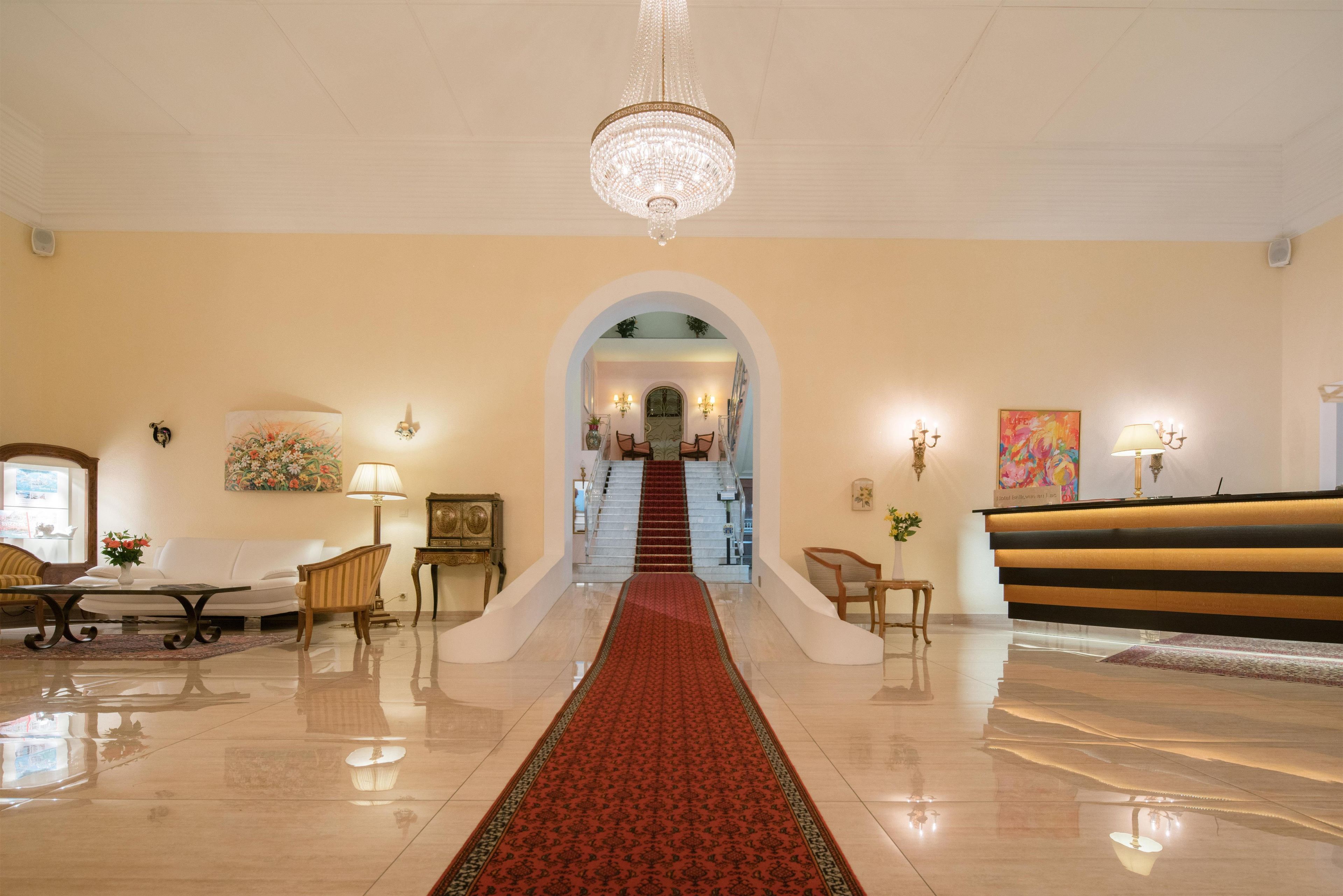 A hotel lobby with a red carpet, a chandelier, a reception desk, and a staircase leading to another floor.