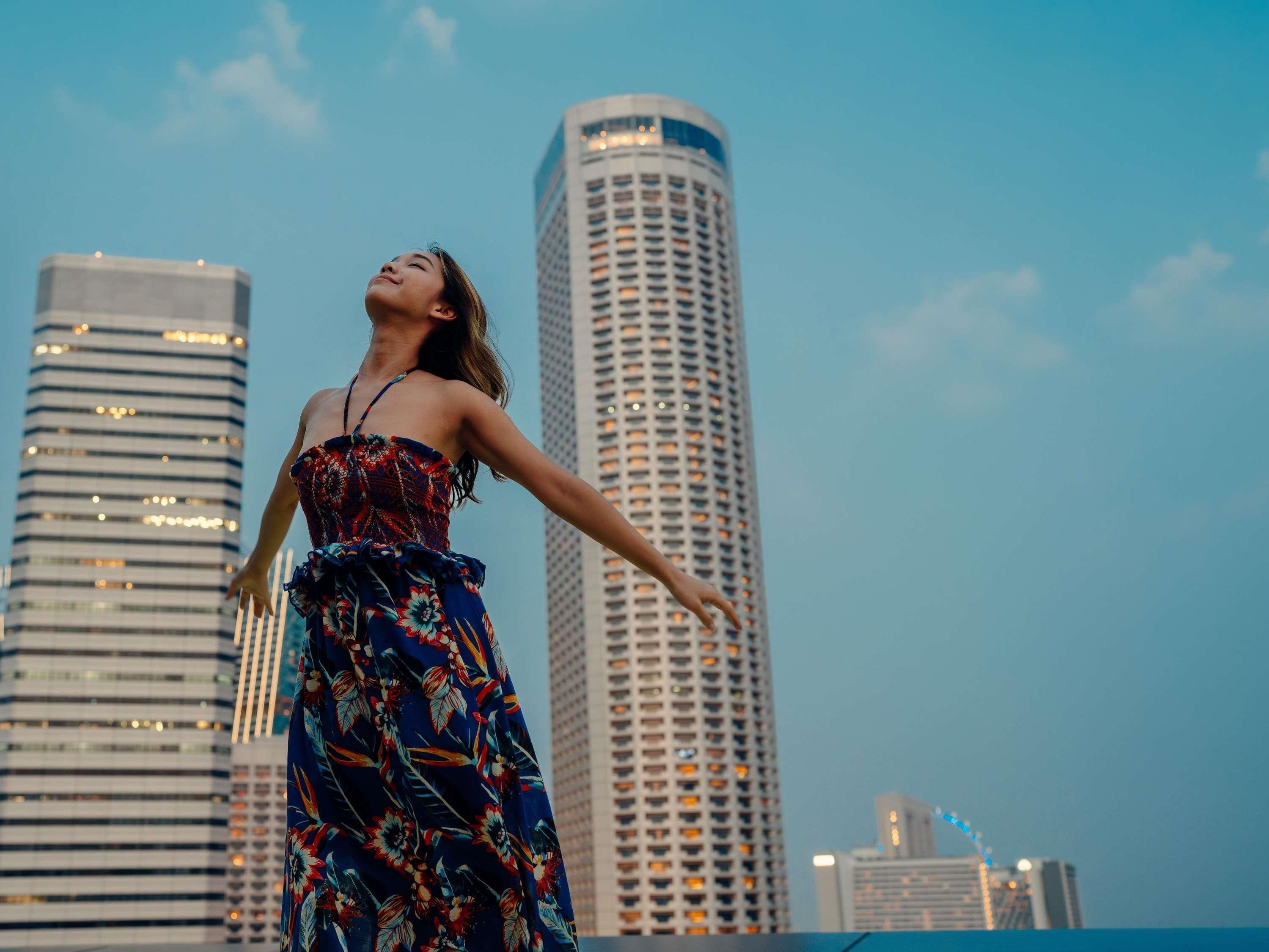 Une personne vêtue d’une robe à motifs floraux, debout, les bras tendus, devant le panorama d’une ville.