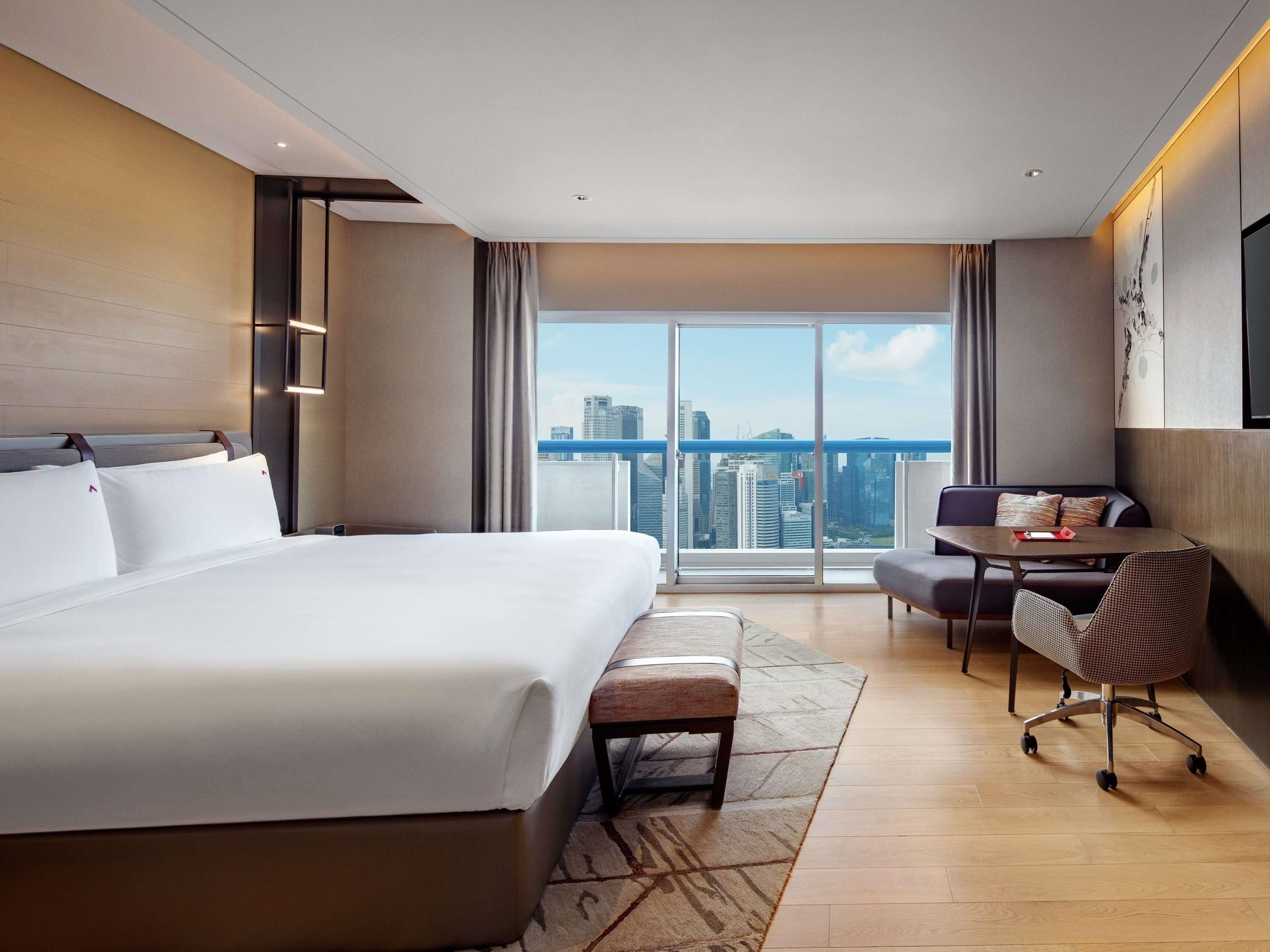 Een hotelkamer met een groot bed, een bureau, een stoel en uitzicht op de stad door grote ramen.