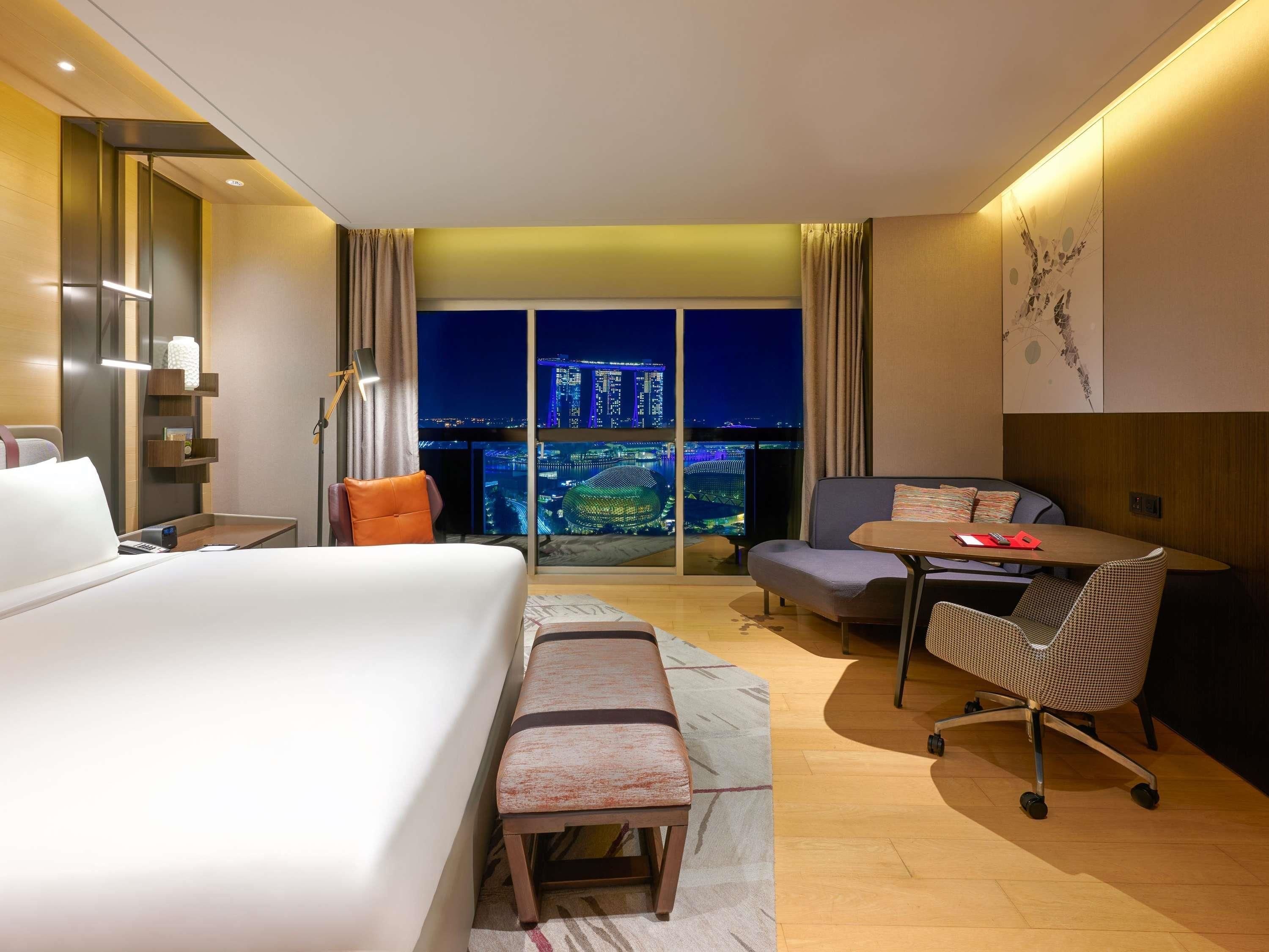 A hotel room with a bed, a desk, a chair, a sofa, and a view of a cityscape at night.