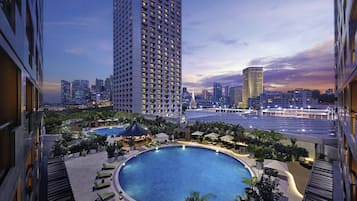 A rooftop pool with lounge chairs, surrounded by greenery, and a city skyline in the background.