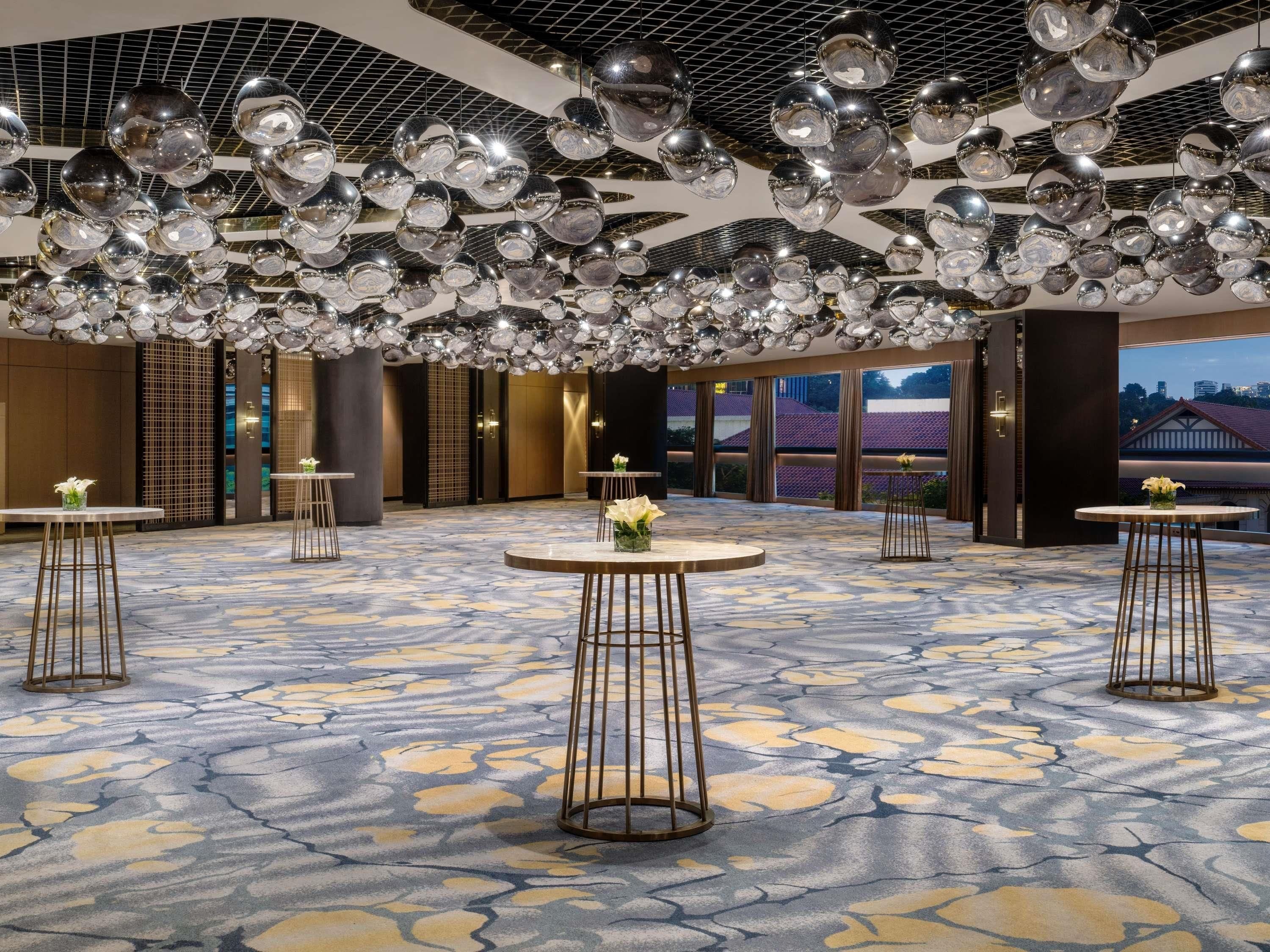 A spacious hotel lobby with a patterned floor, multiple round tables, and a ceiling adorned with reflective spherical decorations.
