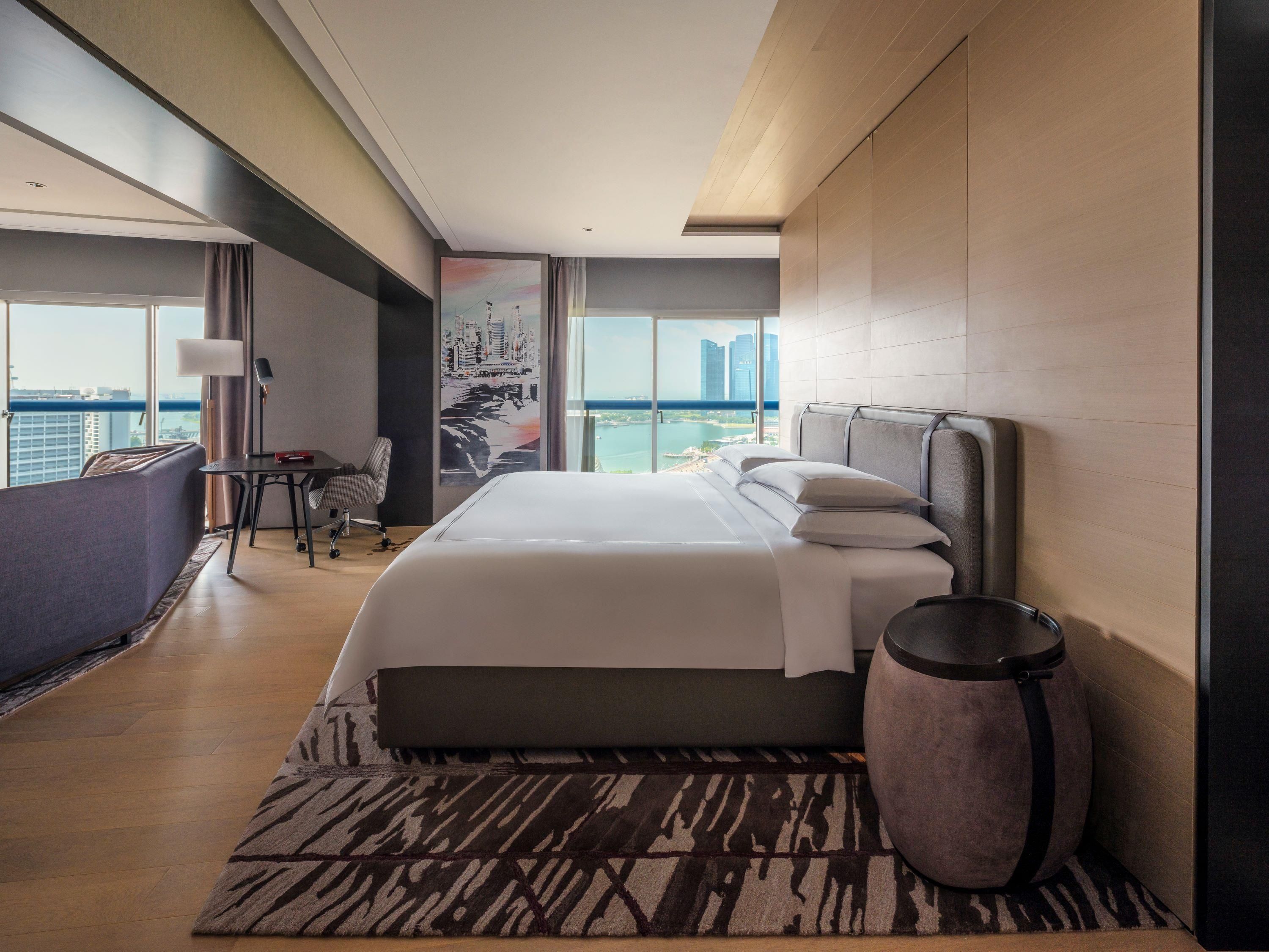 Habitación de hotel con cama, escritorio, silla, ventana grande con vista a la ciudad y un adorno en la pared.