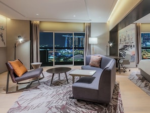 Habitación de hotel moderna con un ventanal grande que ofrece vistas a la ciudad, un sofá, una silla, una mesita y una lámpara.